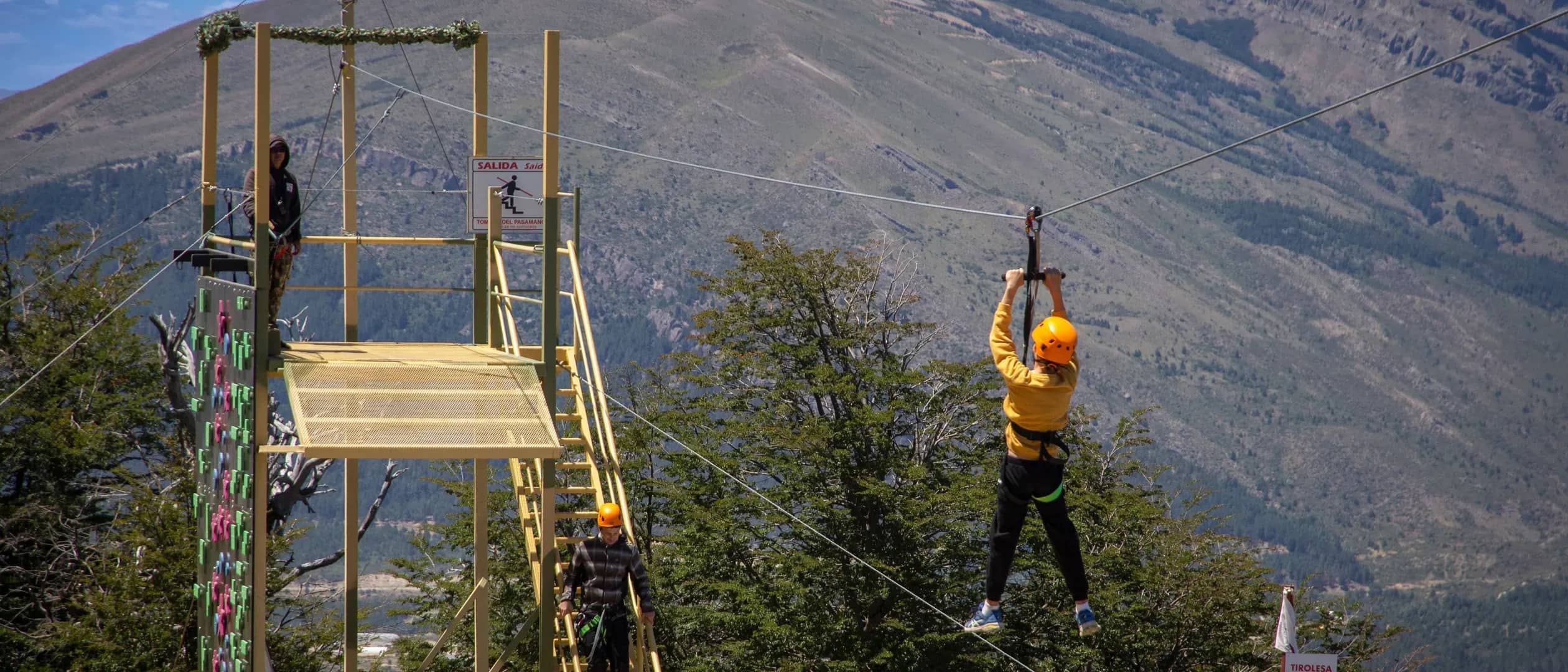 Persona vista desde atrás lanzadose desde la tirolesa con una montaña de fondo en un dia soleado