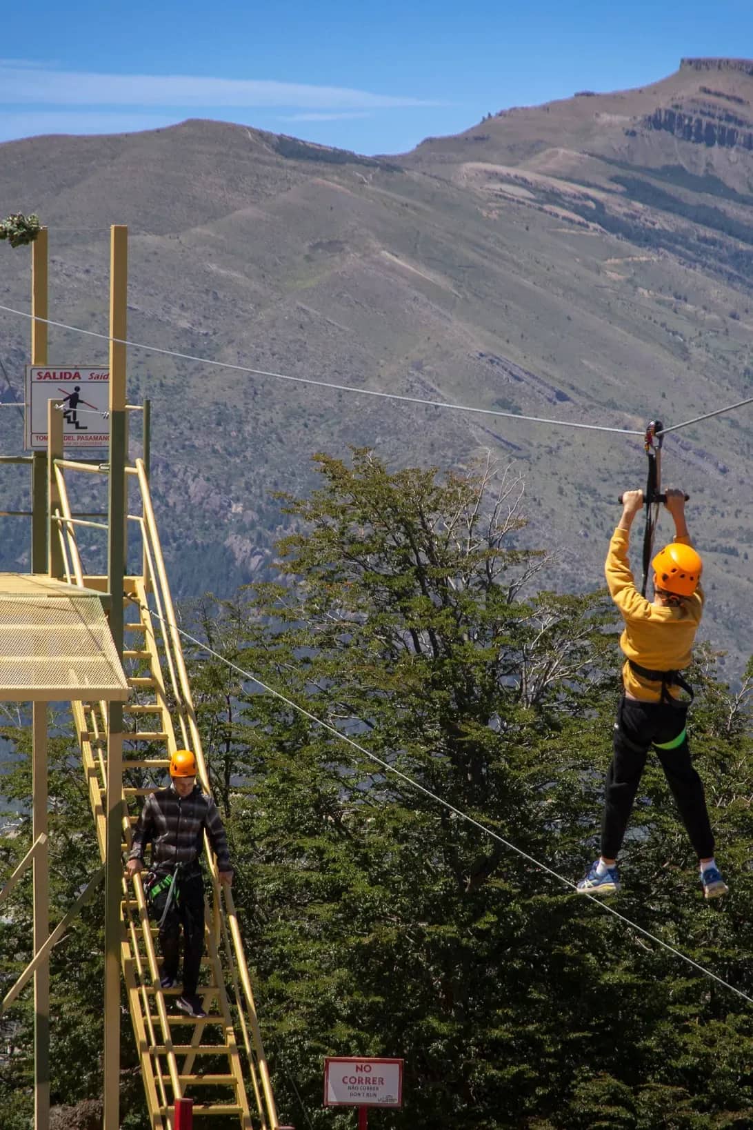Persona vista desde atrás lanzadose desde la tirolesa con una montaña de fondo en un dia soleado