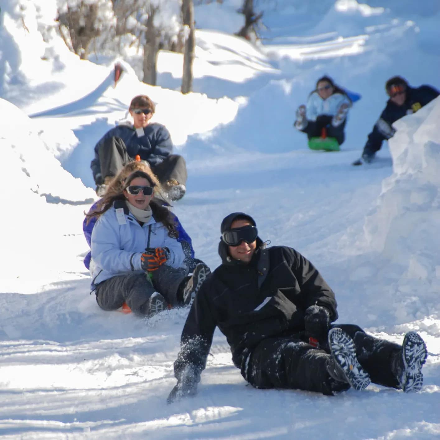 Un grupo de amigos bajando por una pista de nieve en trineos individuales, riendo y expresando gran emoción, con equipos de nieve y cascos en un día soleado de invierno.